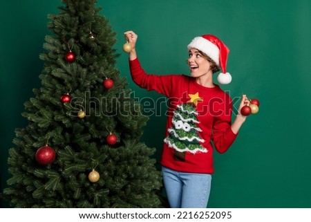 Photo of excited pretty lady wear red sweater headwear hanging tree decoratons isolated green color background
