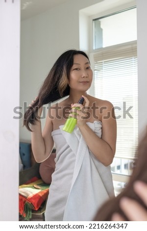 Haircare. Smiling Asian Woman Applying Hair Spray To Split Ends, Young Happy Female Standing Wrapped In Towel After Shower Using avocado conditioner At Home. Royalty-Free Stock Photo #2216264347