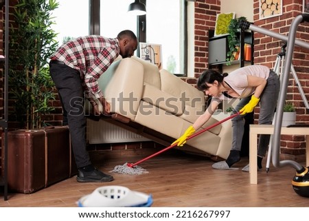 Modern wife mopping under sofa while husband lifts it, multiracial couple doing spring cleaning. Woman and african american man working together to maintain order and cleanliness at home.