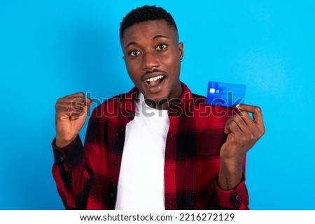 Photo of lucky impressed young handsome man wearing plaid shirt over blue background arm fist holding credit card. Celebrated