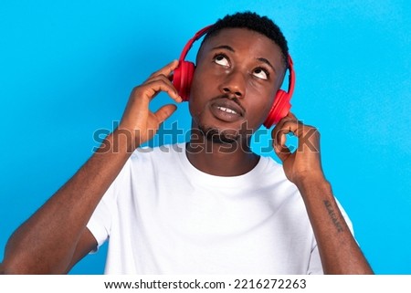 Joyful young handsome man wearing white  T-shirt over blue background sings song keeps hand near mouth as if microphone listens favorite playlist via headphones