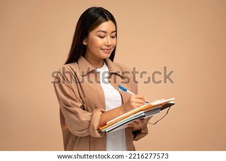 Thoughtful woman standing with paper notebook and pen, making to do list. Indoor studio shot isolated on beige background Royalty-Free Stock Photo #2216277573