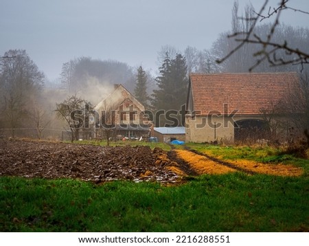 plowed field covered with a snow and old farmhouse on a background in an old village in a rainy misty winter day