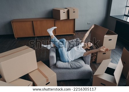 Joyful female homeowner having fun in armchair lifting hands celebrates relocation to new home in room with cardboard boxes. Happy woman tenant feels success on moving day. Mortgage, first realty. Royalty-Free Stock Photo #2216305117