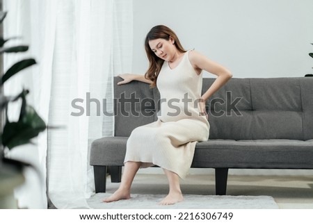 Full body of a beautiful pregnant asian woman is sitting on a couch in the living room, having a back pain as she carries the baby in her tummy. Pregnant problem. Royalty-Free Stock Photo #2216306749