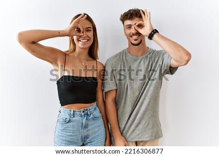 Young beautiful couple standing together over isolated background doing ok gesture with hand smiling, eye looking through fingers with happy face. 