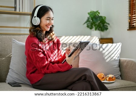 Cheerful and beautiful young Asian female in red sweater, wearing headphones, using her digital tablet, sitting on sofa in living room.