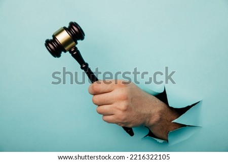 Hand holding a judge's gavel through torn blue paper wall. Law and courts, triumph of justice