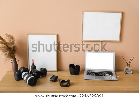 Photographer's workplace with laptop, camera, vase and blank frames near beige wall