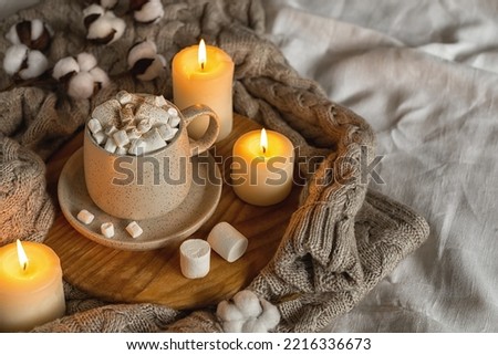 Cozy home still life: hot chocolate or cocoa with marshmallows on a wooden board, a wool sweater, candles and a sprig of cotton. Horizontal photo.The concept of wishing a cozy evening. Selective focus