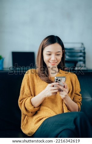 Photo of modern asian woman wearing headphones holding cell phone while lying on sofa in bright apartment