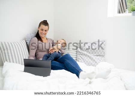 pretty young mother sitting, lying in bed, working on notebook and holding her sleeping baby in her arms and is happy