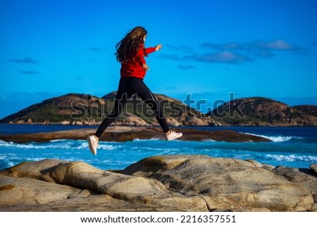 beautiful long-haired girl jumps over rocks by the ocean; girl captured while jumping in motion; sunset on lucky bay beach in western australia