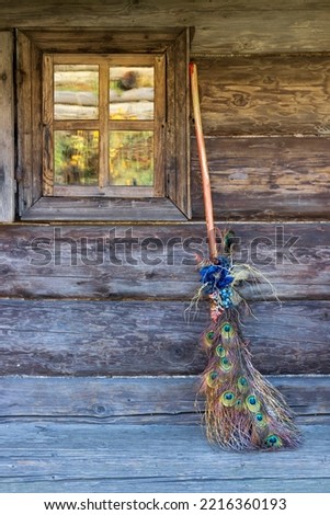 Decorative witch's broom decorated with peacock feathers on the background of a wooden hut with a window. Witch's house. Vertical crop. Copy space Royalty-Free Stock Photo #2216360193