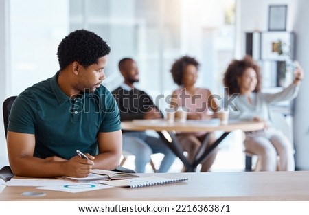Lonely, alone and African businessman in the office working on a project while colleagues take selfie. Reject, sad and antisocial professional employee sitting at his desk as outcast in the workplace Royalty-Free Stock Photo #2216363871
