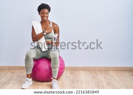 African american woman wearing sportswear sitting on pilates ball cheerful with a smile on face pointing with hand and finger up to the side with happy and natural expression 