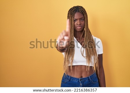 African american woman with braided hair standing over yellow background pointing with finger up and angry expression, showing no gesture 