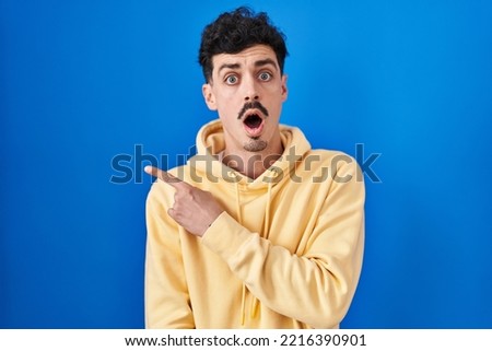 Hispanic man standing over blue background surprised pointing with finger to the side, open mouth amazed expression. 