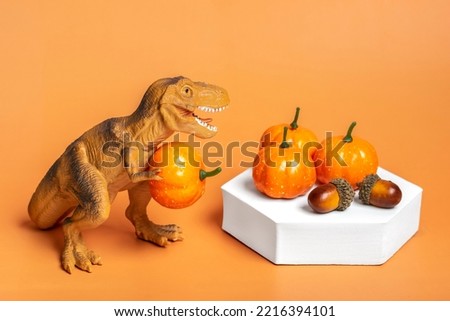 Toy dinosaur Tyrannosaurus holding pumpkin, acorn in its paws, on white podium isolated on orange background Holiday greeting card Happy Halloween day, Hello Autumn creative minimal concept 