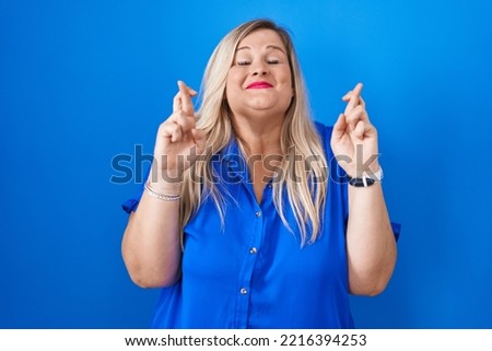 Caucasian plus size woman standing over blue background gesturing finger crossed smiling with hope and eyes closed. luck and superstitious concept. 
