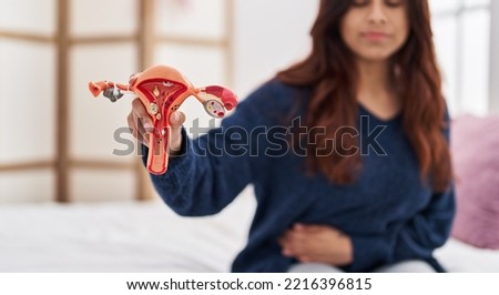 Young hispanic woman suffering for menstrual pain holding anatomical model of fallopian tube at bedroom Royalty-Free Stock Photo #2216396815