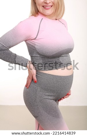 Cropped image of blonde unrecognizable pregnant woman in punk grey tight sports suit, posing over white wall. Simple minimalist background. Studio vertical image with copy space