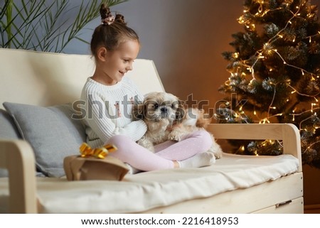 Image of smiling charming little girl with two hair buns wearing casual clothing sitting on sofa with dog, expressing positive emotions, Christmas celebrations.