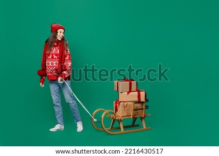 Full size merry young woman wear red warm cozy knitted sweater hat posing carry present box with gift ribbon bow on sled go isolated on plain dark green background Happy New Year 2023 holiday concept