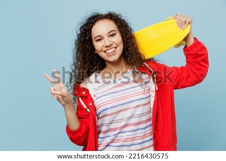 Young woman of African American ethnicity 20s wear red jacket hold skateboard pennyboard behind neck show v-sign isolated on plain pastel light blue cyan background. Wet fall weather season concept