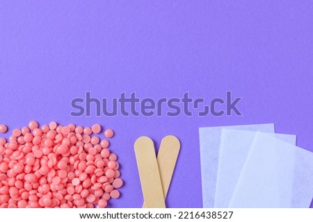 Pink granules of wax for depilation, wooden spatulas and depilatory strips on a lilac background. Epilation, depilation, waxing, unwanted hair removal procedure. 
Place for text.
