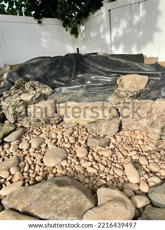 The top down, close up view of a backyard pondless waterfall being installed. The black liner is down and decorative rocks are strategically placed.