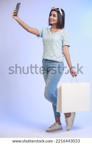 Portrait of happy smiling Asian woman in summer dress holding shopping bag with purchases. Selfie after shopping on mobile phone isolated on a gradient blue background - Copy space for advertisement