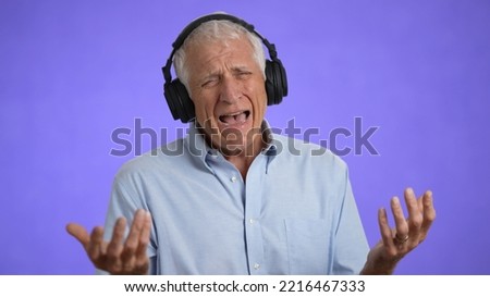 Happy elderly gray-haired man 70s in blue shirt listen music in headphones dance fooling around have fun enjoy relax isolated on plain light purple background studio portrait