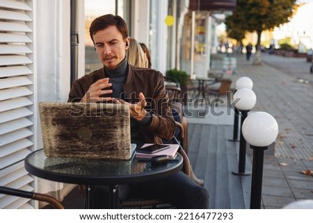 Young male freelancer in wireless earphones having video call and talking during online meeting via laptop in outdoor cafe in city street 