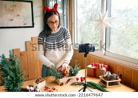 Female florist blogger making winter ikebana with fresh pine branches, candle and christmas decorations in streaming in front of her smartphone. Small business. Seasonal winter workshop.