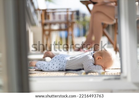 Cute little infant baby boy sulking while playing with toys outdoors at the patio in summer being supervised by her mother seen in the background.