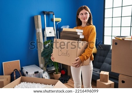 Young brunette woman moving to a new home holding box smiling looking to the side and staring away thinking. 