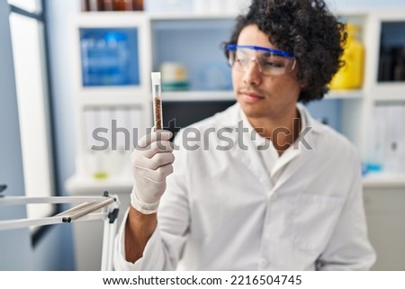 Young hispanic man wearing scientist uniform holding clipboard and test tube at laboratory