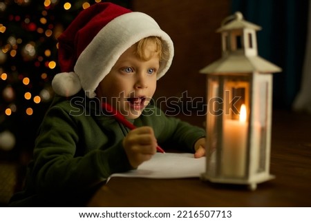 Boy in a Christmas hat looks with shining eyes at a lantern with a candle and dreams of gifts. Child with blue eyes writes a letter on the background of a Christmas tree decorated with a garland