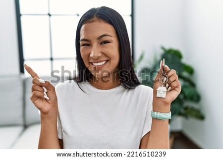Young hispanic woman holding keys of new home with a big smile on face, pointing with hand finger to the side looking at the camera. 