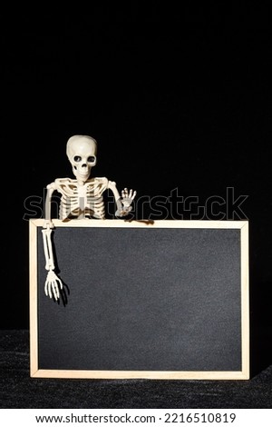 Funny human skeleton holding an empty black blank on a black background, copy space, mockup, vertical frame. Halloween Greeting Card
