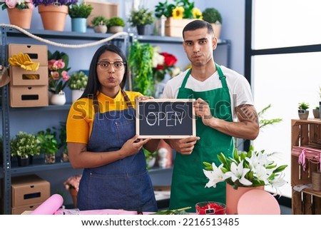 Young hispanic couple working at florist with open sign puffing cheeks with funny face. mouth inflated with air, catching air. 