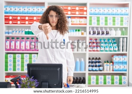 Hispanic woman with curly hair working at pharmacy drugstore looking unhappy and angry showing rejection and negative with thumbs down gesture. bad expression.  Royalty-Free Stock Photo #2216514995