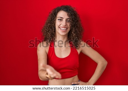 Hispanic woman with curly hair standing over red background smiling cheerful offering palm hand giving assistance and acceptance. 