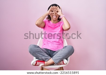 Hispanic young woman sitting on chair over pink background doing ok gesture like binoculars sticking tongue out, eyes looking through fingers. crazy expression. 