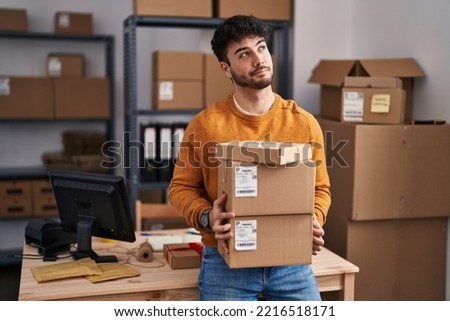 Hispanic man with beard working at small business ecommerce holding packages smiling looking to the side and staring away thinking. 
