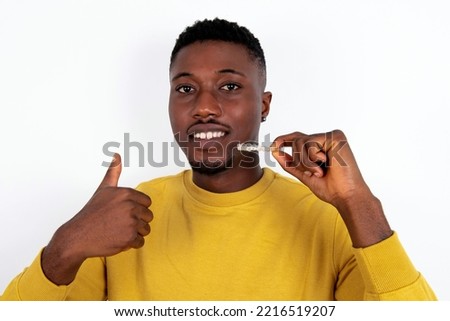 young handsome man wearing yellow sweater over white  background holding an invisible braces aligner and rising thumb up, recommending this new treatment. Dental healthcare concept.