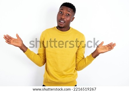 So what? Portrait of arrogant young handsome man wearing yellow sweater over white  background shrugging hands sideways smiling gasping indifferent, telling something obvious. Royalty-Free Stock Photo #2216519267