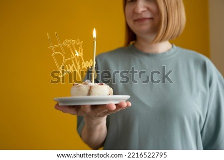 A happy woman holds a plate with homemade cakes and a candle on a yellow background, a happy Birthday topper in her hands.