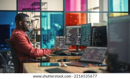 African American Man Writing Lines Of Code On Desktop Computer With Multiple Monitors and Laptop in Creative Office. Male Data Scientist Working on Innovative Online Service For Start-up Company. Royalty-Free Stock Photo #2216535677